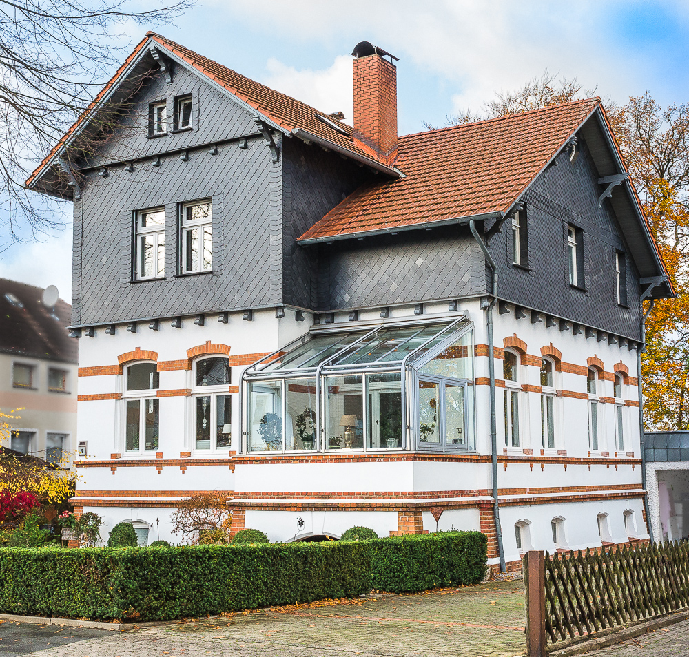 Ein historisches Gebäude mit einem Glaswintergarten und einem roten Ziegeldach in einer städtischen Umgebung.